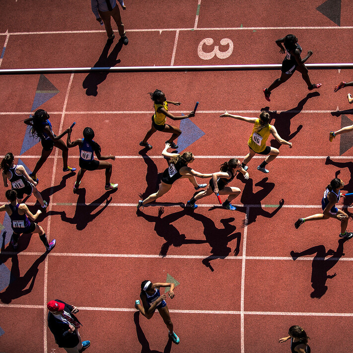 An aerial view of a crowd of rushing sprinters handing off batons in a relay race